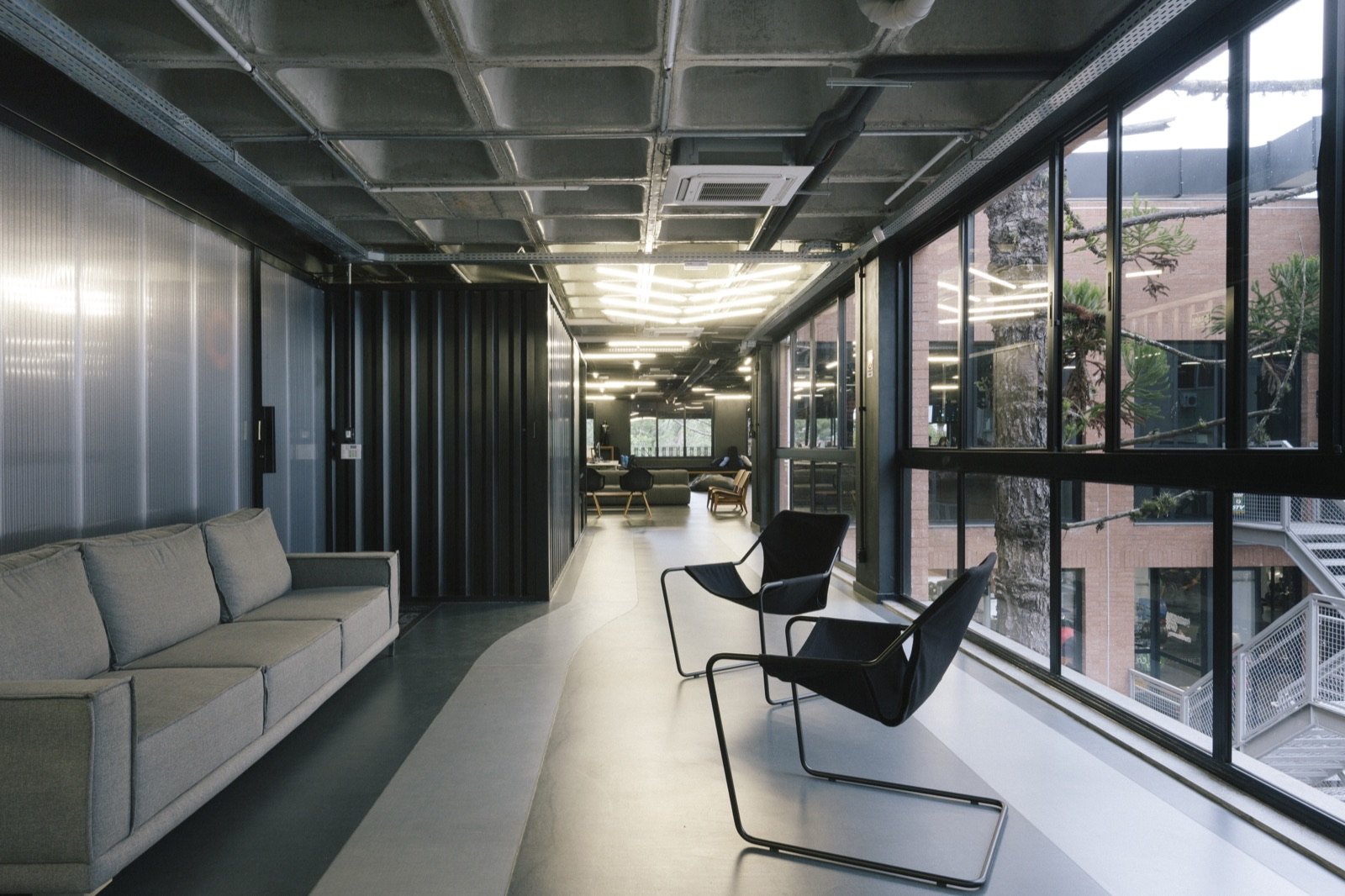 A lounge corridor with a sofa and two sling chairs along a wall of windows