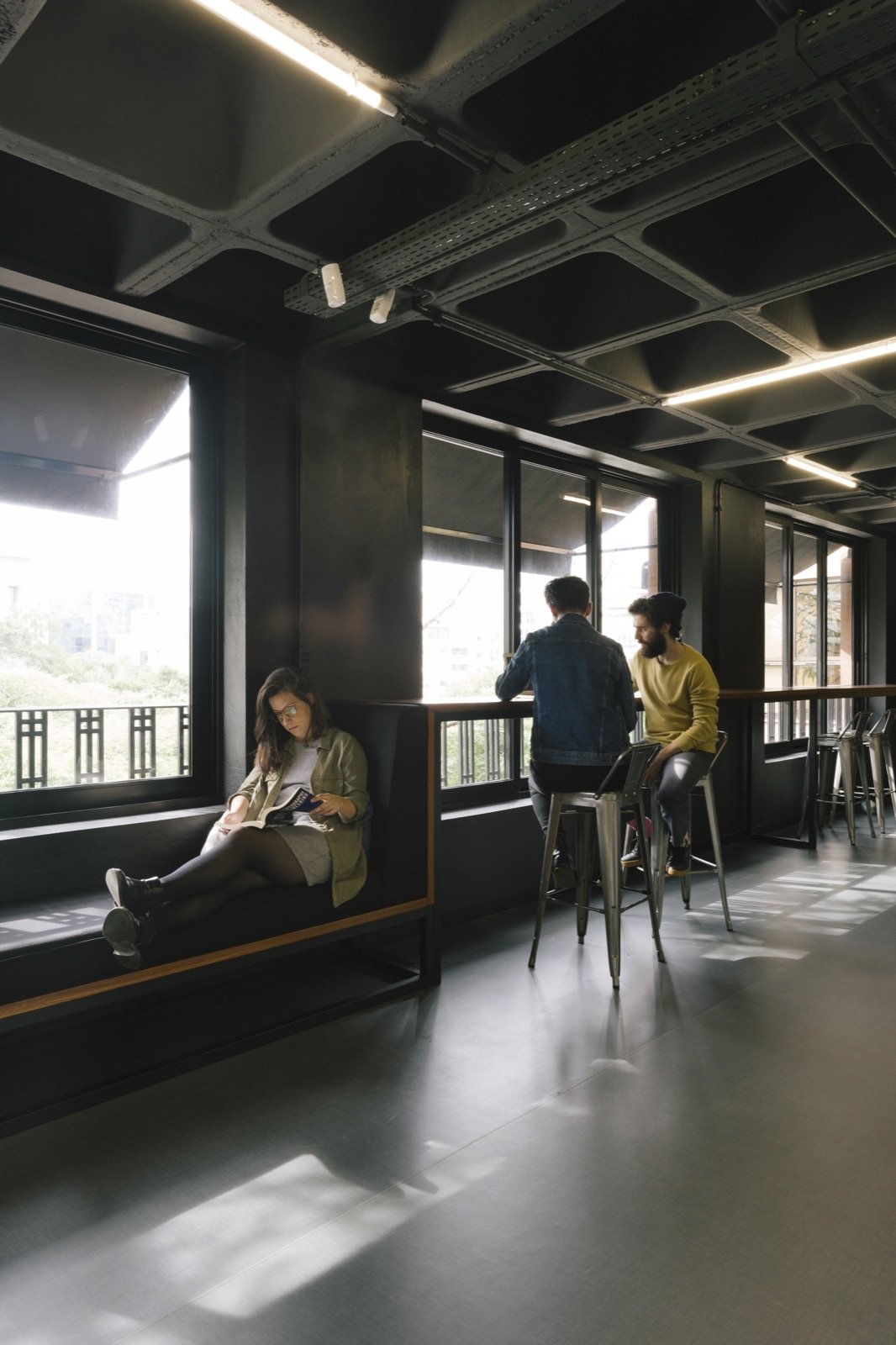 One person reading on a window bench while two colleagues talk at a window counter
